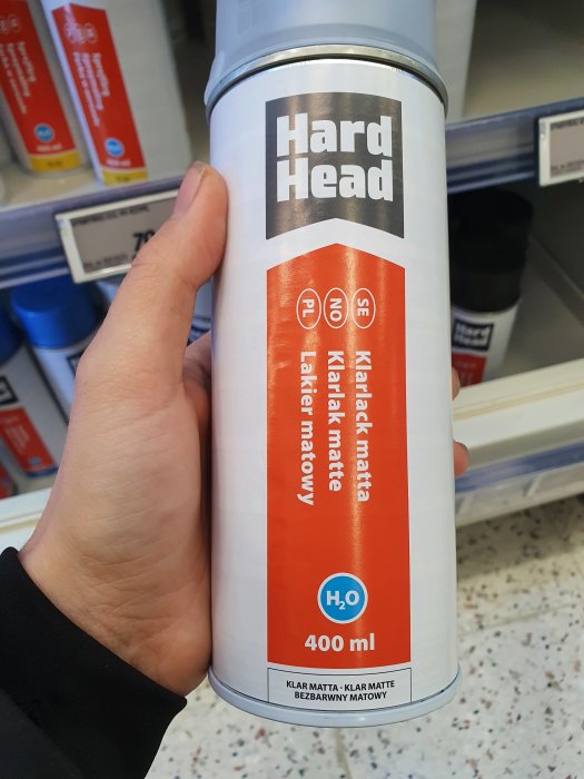 Hand håller en sprayburk med klar matt lack från Hard Head i en butikshylla.