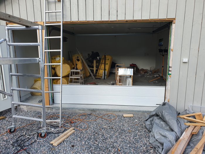 Garage under renovering med öppen port, byggmaterial och stegar inuti samt på grusgolvet framför.