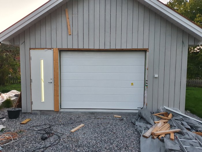Nybyggt garage i skymningen med stängd port, en tänd utomhuslampa, ojämnt fördelade brädor och byggmaterial på grusgolvet.