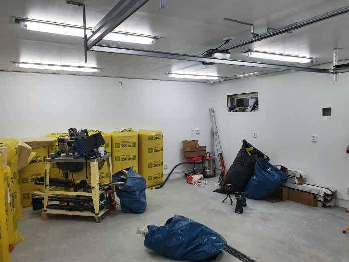 Interiör av ett nyrenoverat garage med vitmålade väggar, infällda taklampor och byggmaterial inklusive gul isoleringsskivor.