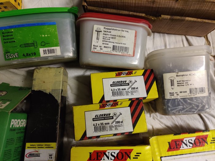 Olika förpackningar med skruvar, inklusive trälåda och plastburkar, etiketter synliga.