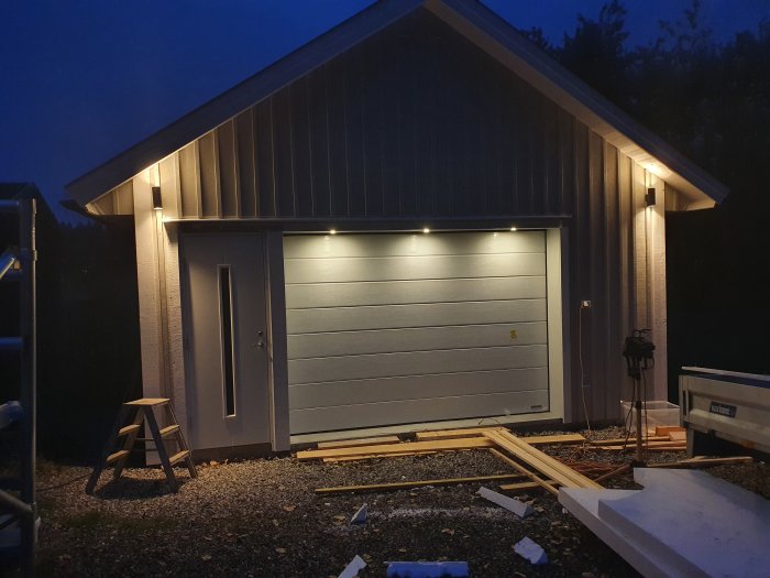 Nyligen renoverat garage i skymningen med öppen dörr, fönster och upplyst med utomhusbelysning, byggmaterial framför byggnaden.