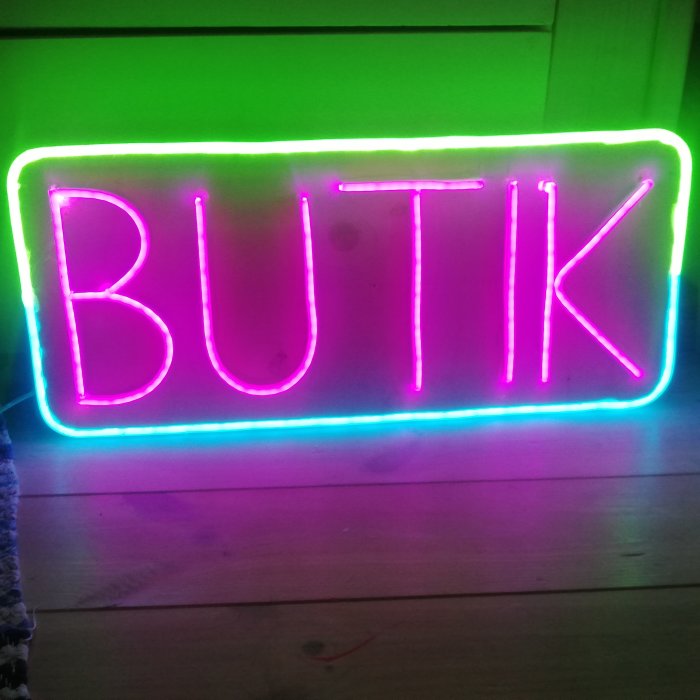 Hemmagjord neonskylt med ordet "BUTIK" i lysande rosa och gröna LED-lister mot en mörk bakgrund.