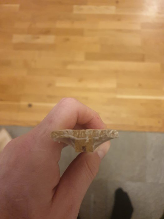 Sprucken tröskellist hålls mellan fingrar framför parkettgolv och stenplattor.