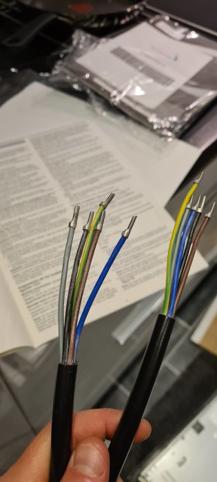Hand som håller två elkablar med blottade ändar framför en oskarp spishäll och installationsmanual.