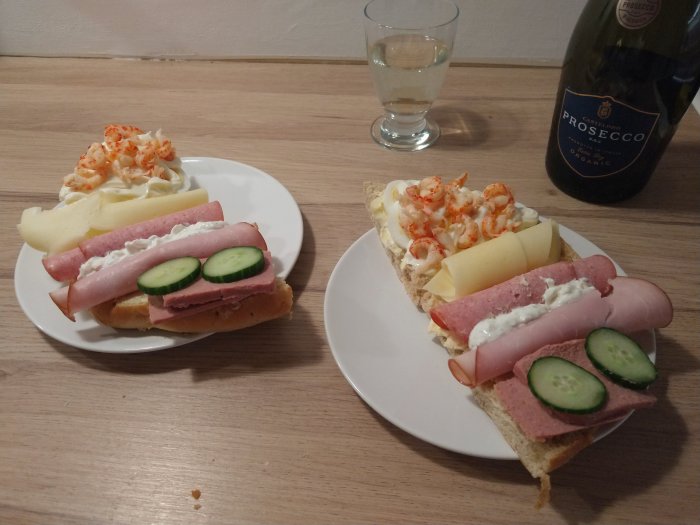 Två smörgåsar med ost, skinka, räkor och gurka på tallrik med ett glas och flaska prosecco.