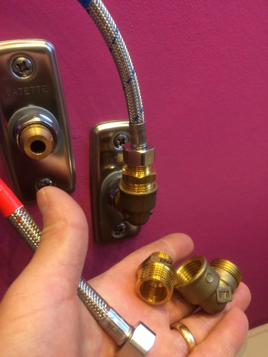 Hand håller VVS-komponenter framför en vägghängd blandare med slangkopplingar och rördelar.