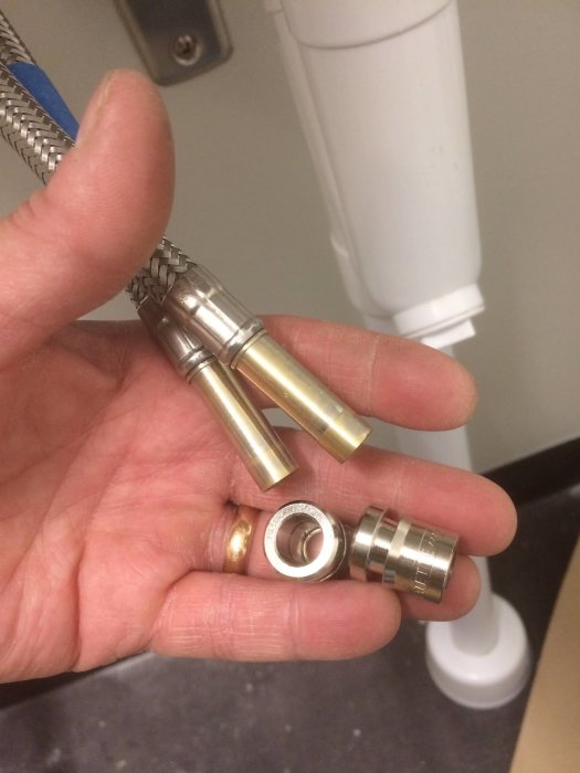 Hand håller olika VVS-kopplingar inklusive blandarmutter, klämkoppling och kulventil framför WC-installation.