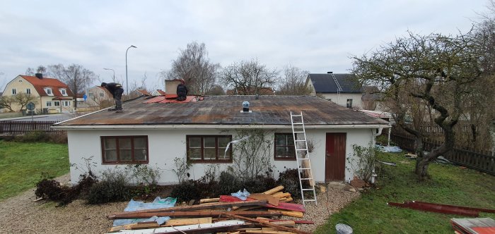 Arbetare river gammal takpapp på ett hus i förberedelse för att lägga ny, med material och stegar runtomkring.