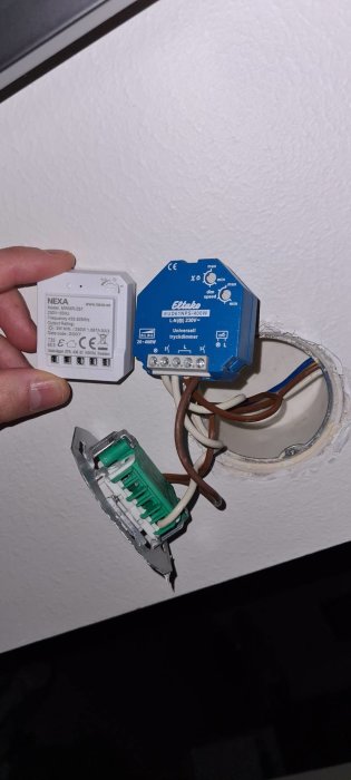 En hand håller en Nexa-mottagare bredvid en öppen Eltako dimmer installerad i vägg med synliga kablar.