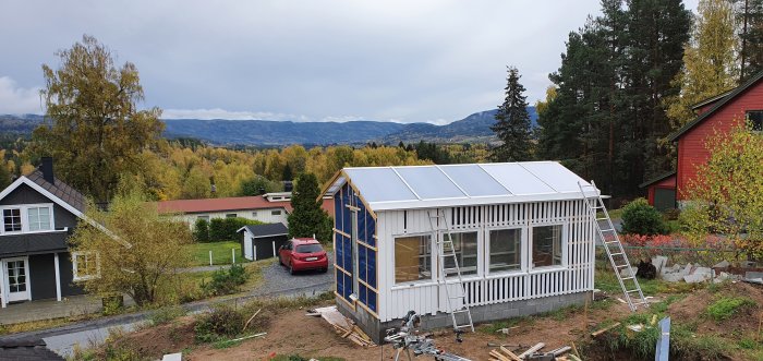 Nybyggt växthus med OSB-panel och vita väggar under konstruktion, i en trädgård med bergig bakgrund.