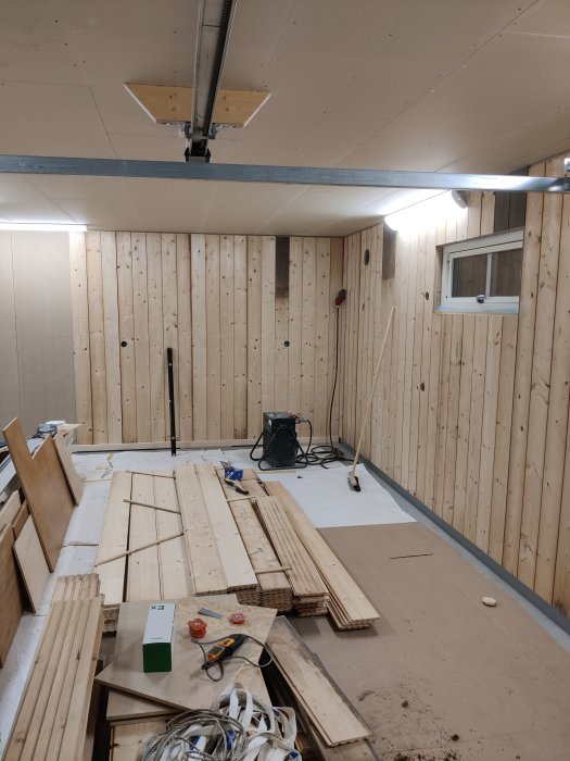 Interiör av en garageverkstad under renovering med träpaneler på väggarna och en samling byggmaterial och verktyg på golvet.