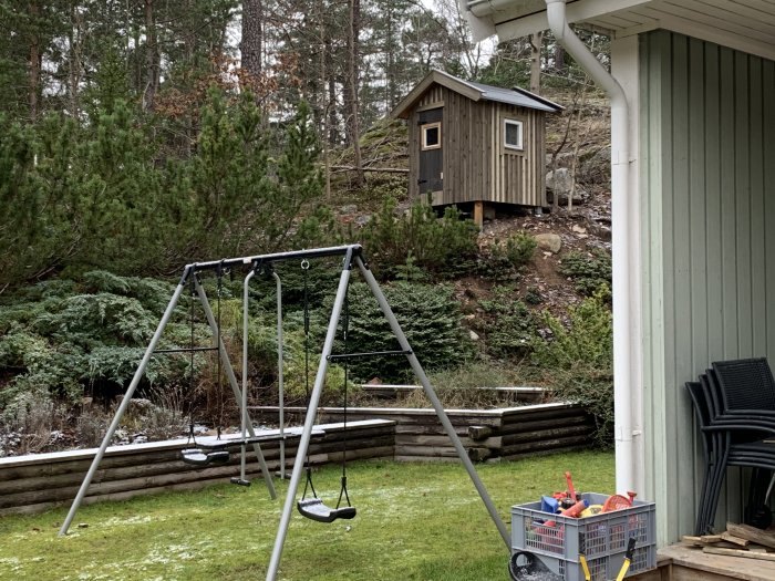 Spräcklig lekstuga på en kulle med trävirke av olika nyanser, synlig från en trädgård med gungställning och verktyg nära hus.