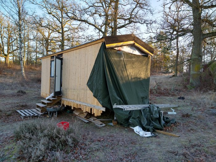 Ofärdigt trähus med överdrag och byggmaterial runtomkring, omgivet av träd och frostig mark.