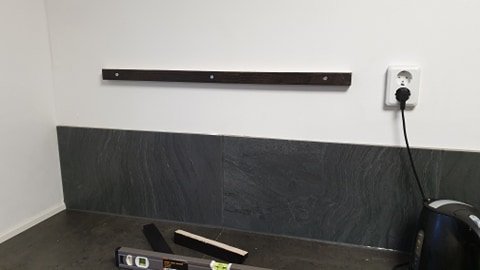 Mörkbetsad stödregel fastskruvad på en ljus vägg över en mörk kaklad yta, med verktyg och delar på golvet.