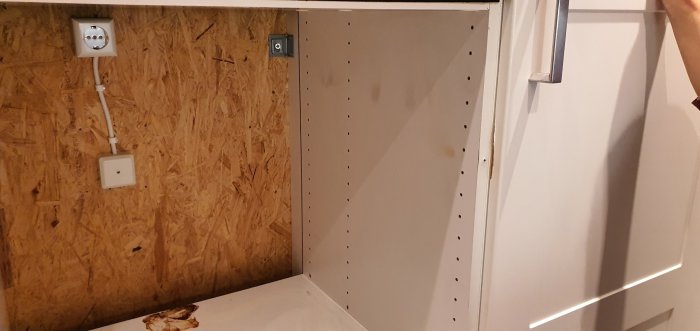 Tomt köksskåp utan ugn med synlig OSB-vägg och eluttag, skåpdörr till höger.