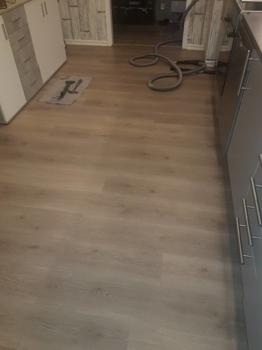 Köksgolv i laminat med en dammsugarslang på golvet och skåp på sidan.
