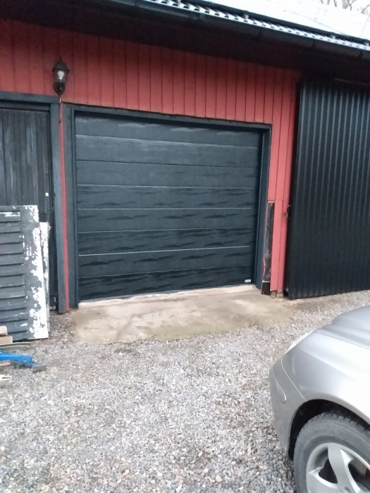 Nyinstallerad svart garageport på rödmålat hus med grundmålat foder och en bilparkering framför.