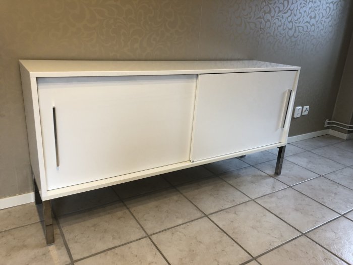 Vit IKEA skänk med två dörrar och metallben, placerad mot en mönstrad vägg och kakelgolv.