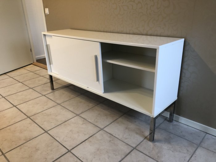 Vit IKEA tv-bänk med öppna hyllor och dold förvaring, på metallben, mot en tapetserad vägg.