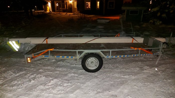 Släpvagn lastad med staplade träplankor av olika längder, säkrade med orange spännband på en snöbelagd yta på kvällen.