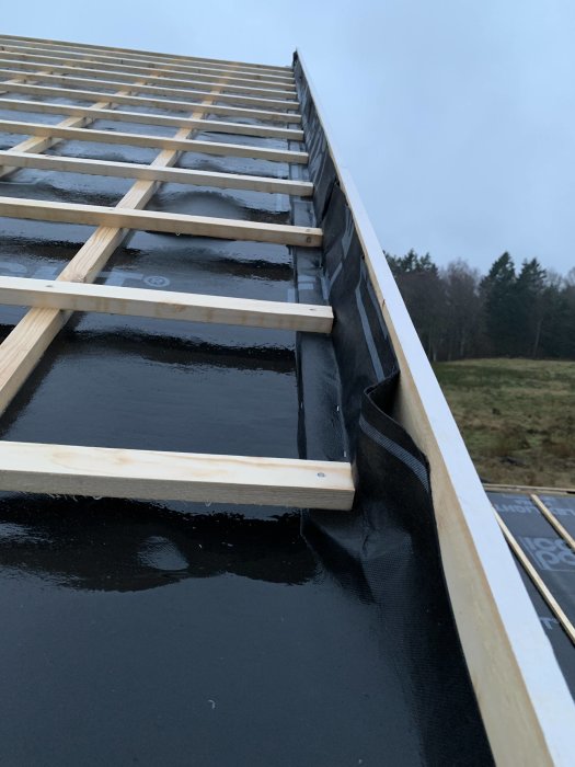 Del av takstomme med svart underlagsduk och placerade träreglar vid dagsljus.