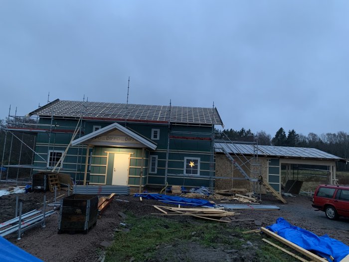 Hus under konstruktion med takstolar, byggnadsställningar och presenningar på byggarbetsplats vid skymning.