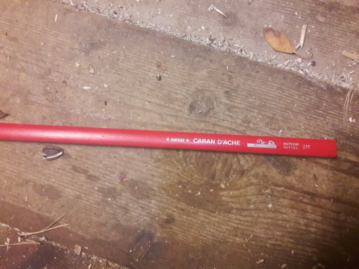 Röd blyertspenna från Caran d'Ache på ett slitet trägolv med spån och små skräp.
