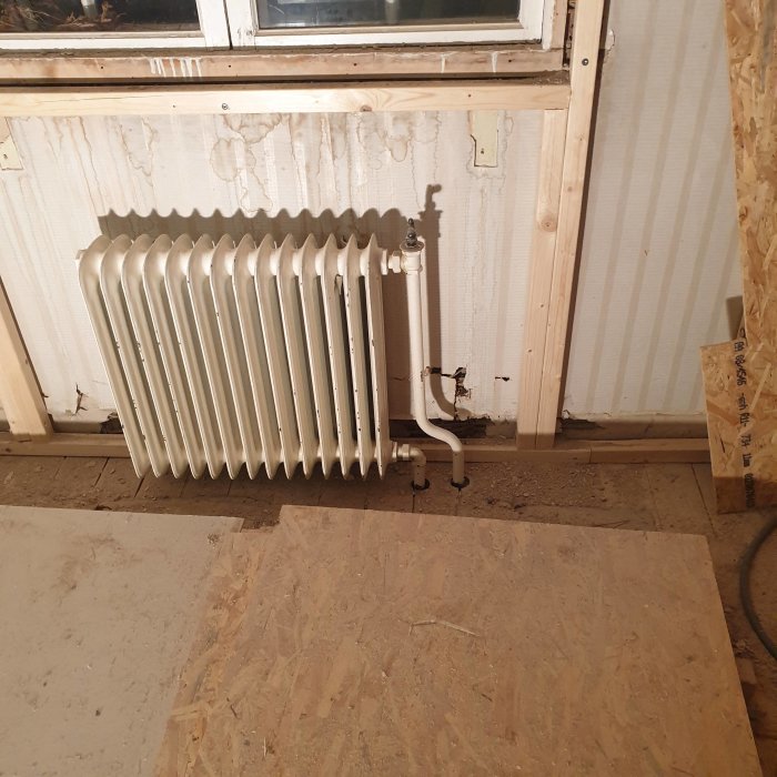 En vit radiator monterad under ett fönster i ett rum under renovering med träreglar och oskyddad isolering.