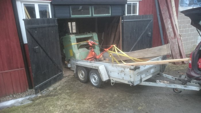 En stor sågmaskin lastad och fastspänd på en släpvagn framför en lada.