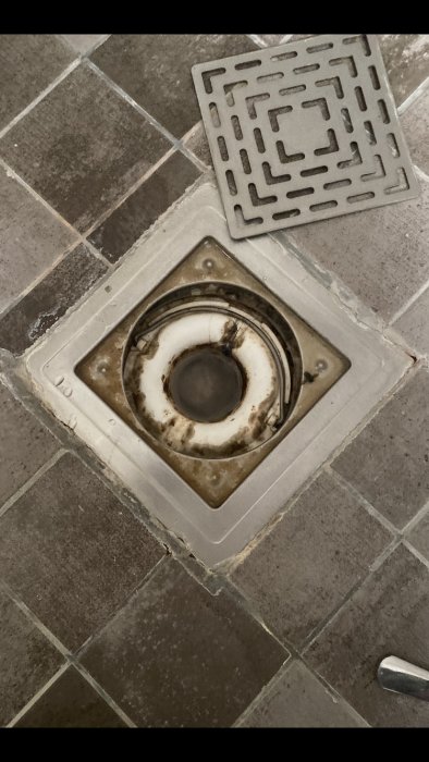 Golvbrunn med borttagen metallrist som visar ackumulerad smuts och kalkavlagringar kring avloppet.