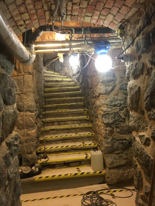 Renoverad stentrappa i källare med gula och svarta varningstejpade steg och exponerat tegeltak.