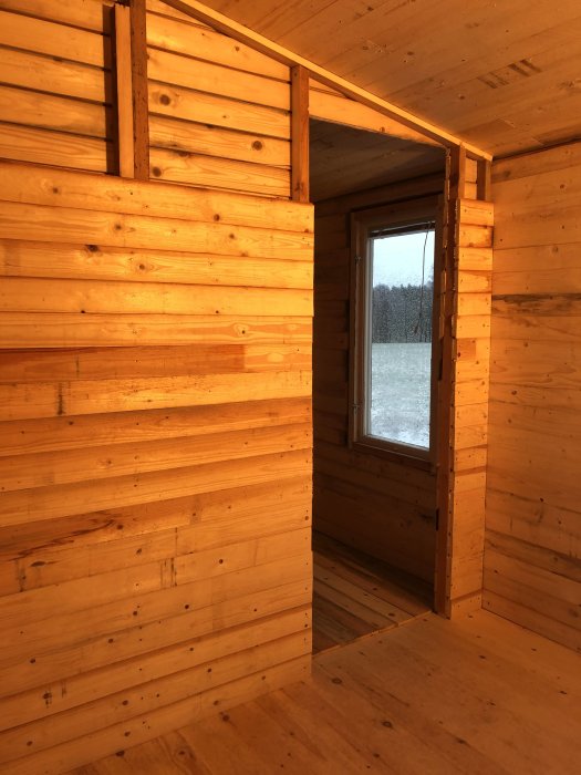 Nybyggd innervägg av råspont i trä med fönster mot vinterlandskap.