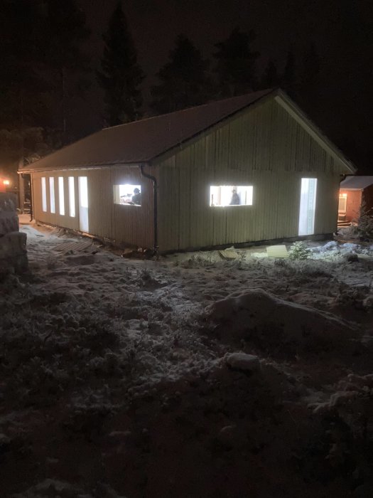 Ett upplyst hus under konstruktion på kvällen med snö på marken.