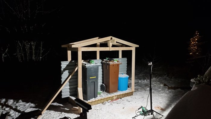 Ett oavslutat litet hus för soptunnor under konstruktion på en trall vid natt.
