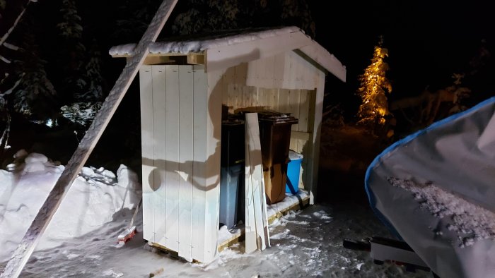 Ett vedskjul i snöig bakgård på natten belyst av yttre ljuskällor med diverse veden och verktyg inne.