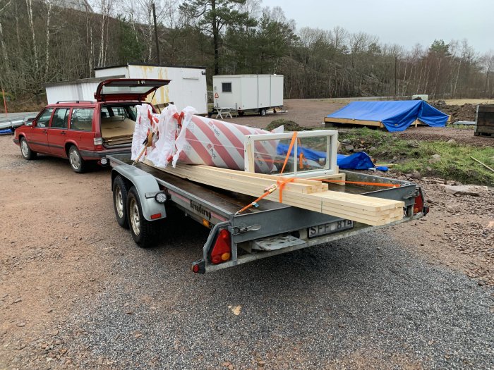 Byggmaterial och isolering fastsäkrade på släpvagn bakom röd kombibil.