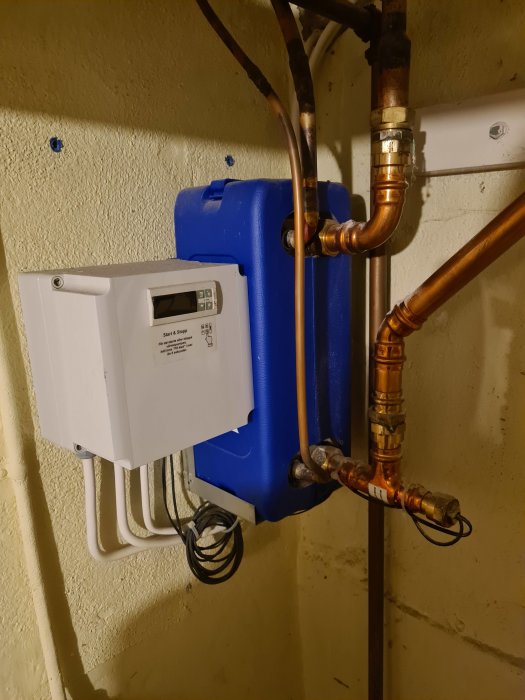 Installation av en vit värmepumpsenhet bredvid en blå tank med kopparledningsanslutningar på en murad vägg.