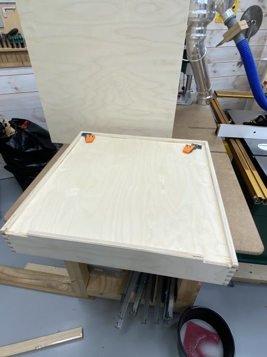 En plywoodlåda under konstruktion med skenor och verktyg synliga i en verkstadsmiljö.