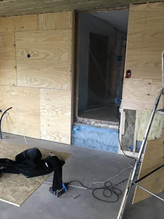 Obehandlat betonggolv och plywoodvägg med öppning för planerad trappa mellan husnivåer.