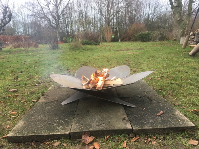 Eldfat av svetsat metall konstruerat med överlappande ben står på marksten med brinnande ved.