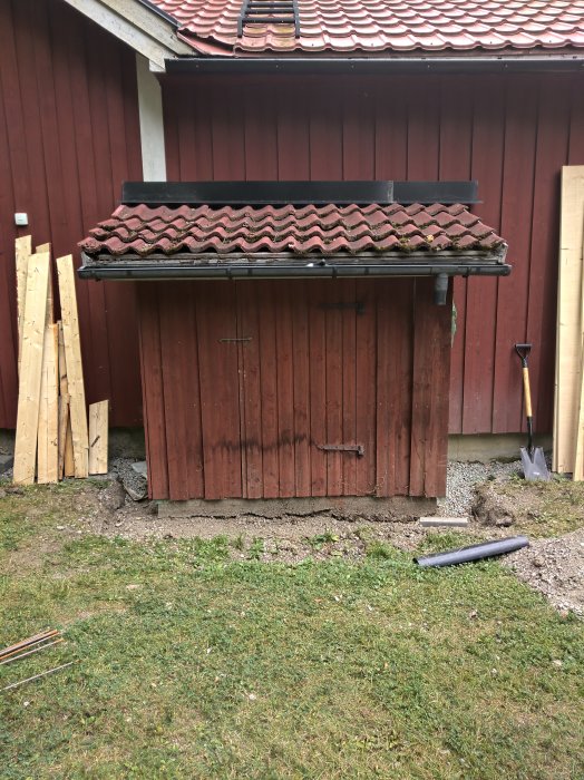 Utgångsläget för renovering: ett litet rött hobbithus med tegeltak, omgivet av byggmaterial och spade.