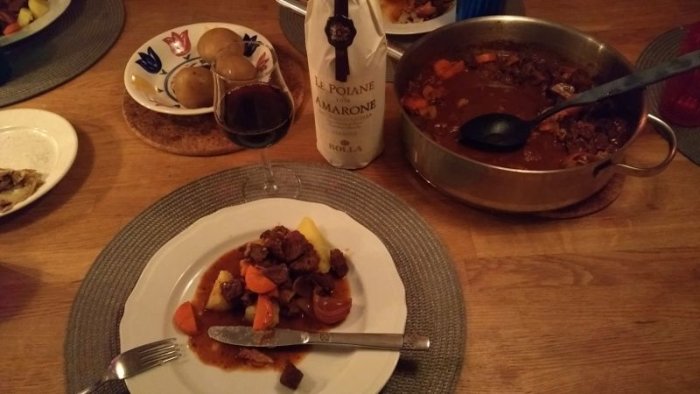 En middagsdukning med en tallrik gryta, glas vin och en flaska, samt en gryta på bordet.