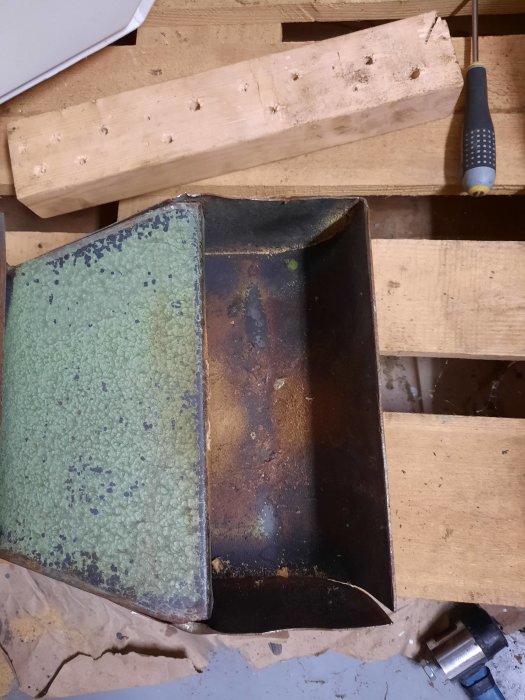 Metallremskydd med bananform som rätats ut, placerat på ett arbetshyvelbänk, bredvid en gummimatta och verktyg.