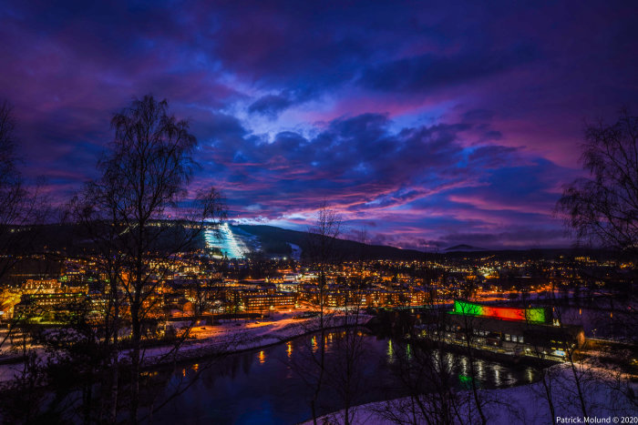 Kvällsvy med upplyst skidbacke över snötäckt stad med flod och dramatisk himmel.