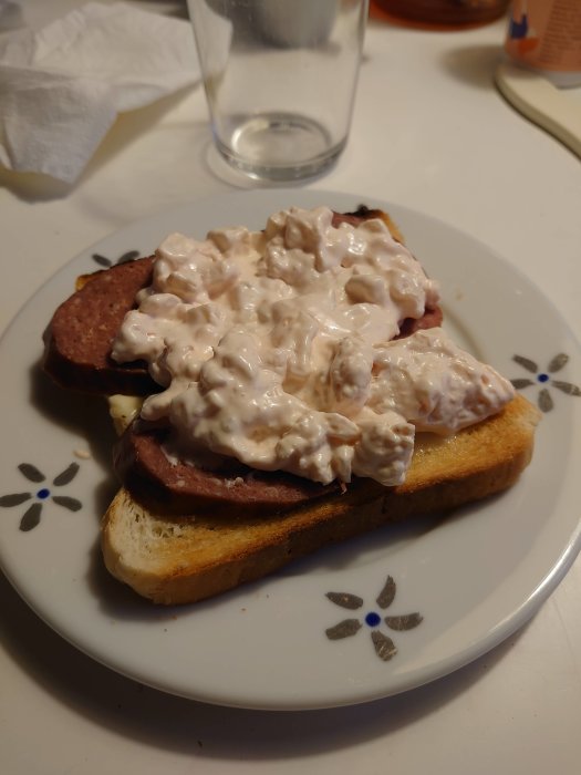 Macka med leverpastej och skagenröra på en vit blommönstrad tallrik, bredvid ett tomt glas och en servett.