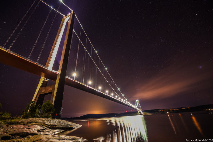 Högakustenbron belyses av lampor i gryningen med stjärnhimmel och spegling i vattnet.