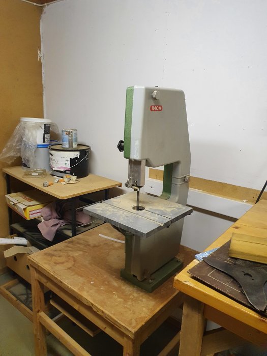 Äldre Inca bandsåg på arbetsbänk i verkstad, med tillbehör och färgburkar i bakgrunden.