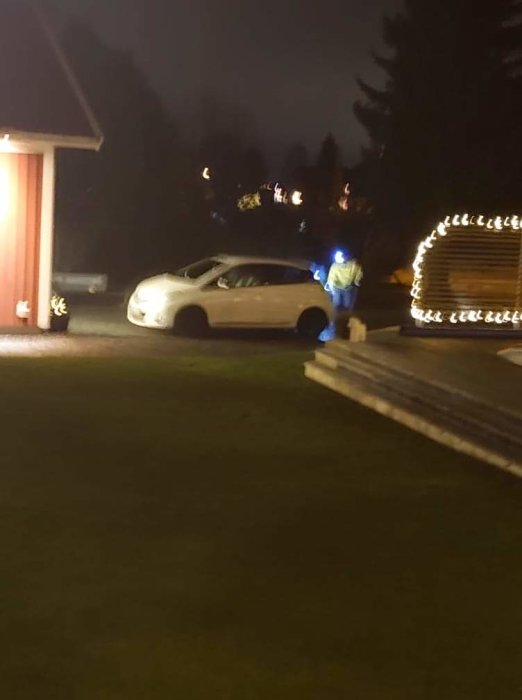 Person med pannlampa tvättar bil i mörker vid uppfart nära carport med ljusdekorationer.