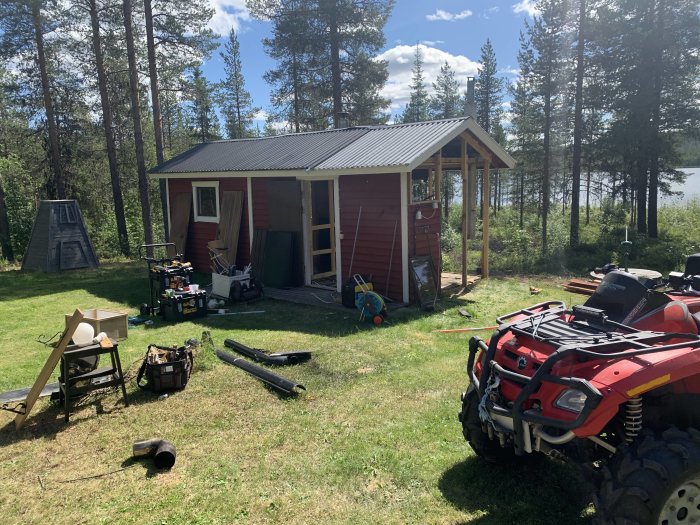 Arbetsplats utomhus med röd liten byggnad under rivning, diverse byggmaterial och verktyg utspridda och en fyrhjuling i förgrunden.